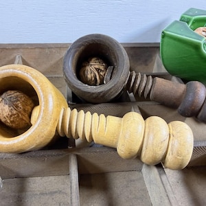 May include: Two wooden nutcrackers with screw mechanisms, one light tan and one dark brown, each holding a whole walnut. The nutcrackers are positioned on a wooden grid surface next to a green ceramic dish containing walnut shells.