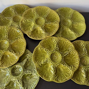 May include: Seven olive green ceramic oyster plates are displayed. Each plate has six compartments around a central well, with a textured, floral design. The plates are arranged on a dark surface. The plates are approximately 25 cm in diameter.