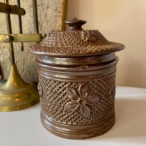 May include: A brown ceramic jar with a lid, featuring a woven pattern and floral designs. The jar has a slightly textured surface and a glossy finish.