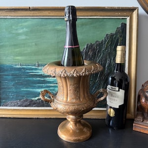 May include: A metallic bronze-toned wine cooler featuring an ornate rim with leaf-like detailing, two side handles, and a pedestal base. The cooler holds a dark glass bottle with a black foil top. A second wine bottle with a white label stands nearby.