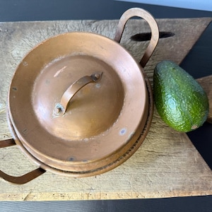 May include: A copper cooking pot with a lid and handle, placed on a wooden chopping board. The pot has a rustic look. A green avocado is next to the pot on the board. The chopping board is uniquely shaped and made of wood.