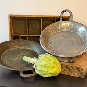 May include: Two antique metal pans with handles, one resting on a wooden block. A wooden crate is behind the pans. A fresh green artichoke is in the foreground. The pans have a rustic, aged appearance.