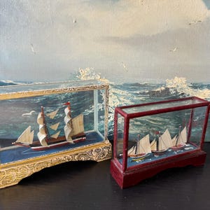 May include: Two model ships enclosed in rectangular glass display cases. The larger ship features white sails and a red hull inside a gold-toned, ornate base. The smaller ship has white sails inside a dark red, smooth-finished wooden base.
