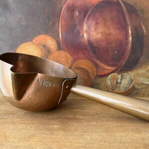 May include: A hammered copper saucepan with a pouring spout and a long, straight metal handle. The exterior of the bowl features a stamped hallmark and the number 12. The vessel has a rounded base and a polished metallic finish throughout.