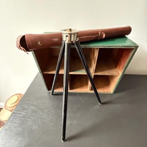 May include: A vintage black and silver tripod with a brown leather case. The tripod has three black legs and a silver top. A brown leather case is positioned behind the tripod, and a green and brown wooden storage unit is in the background. The tripod is on a dark surface.