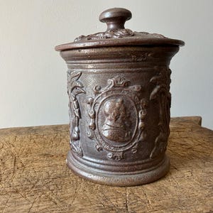 May include: A brown ceramic jar with a lid, featuring ornate relief details. The jar has a cylindrical shape with a slightly flared base and a rounded lid with a small knob. The relief includes framed portraits and floral patterns. The jar is displayed on a wooden surface.