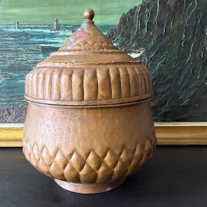 May include: A round, lidded container crafted from hammered copper with a warm, aged patina. The lid features a pointed finial and a textured, diamond-patterned band. The base of the vessel also displays a repeating, carved diamond motif.