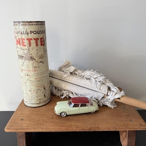 May include: A tall, cylindrical, weathered metal container with the text "AVAL-POUSSIERE" and "NETTE" in red and black, a white cotton string mop with a wooden handle, and a small, pale green toy car with a dark red roof.
