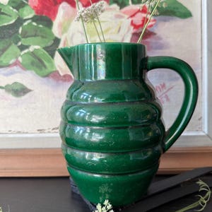 May include: A dark green ceramic jug with a ribbed design. The jug has a handle and a spout, and is filled with white wildflowers. A painting of roses is in the background.