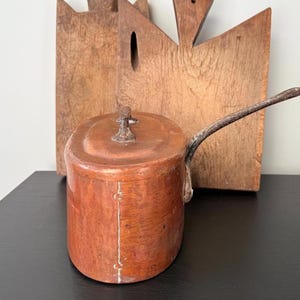 May include: A small, antique copper-coloured pot with a long, curved handle and a lid. The pot has a vertical white line down the front. Two wooden chopping boards are in the background.