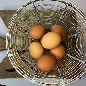 May include: A white metal egg basket with six brown eggs inside. The basket is sitting on a wooden chopping board.