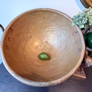 May include: A large, round, tan bowl with two dark brown handles. A small green avocado rests in the centre of the bowl. The bowl has a rustic, handcrafted appearance, suggesting it is made of clay or a similar material.
