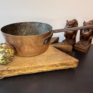 May include: A copper saucepan with a long handle rests on a wooden chopping board. A green and yellow fruit and two brown gargoyle statues are also present. The pan has a dark interior and a riveted handle.