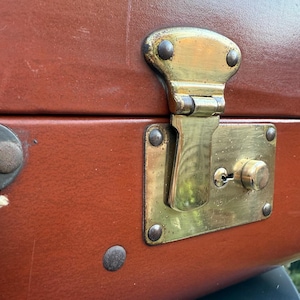May include: Close-up of a brown leather suitcase with a brass lock and keyhole. The lock has a decorative design and is secured with rivets.