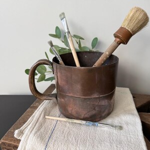 May include: A copper-coloured mug with a handle contains several paint brushes. The brushes have wooden handles and various brush heads. One brush lies on a white cloth. The mug sits on a wooden surface.
