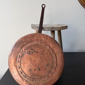 May include: An antique copper pan with a long, riveted handle. The pan features a circular design with a raised rim and rivets around the edge. The copper has a warm, reddish-brown hue, indicating age and use, typical of vintage cookware.