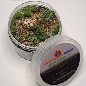 Op de afbeelding: Een doorzichtige plastic container met een deksel. De container is gevuld met een bruin en groen mengsel. Het deksel heeft een label met de tekst "Moonhopper Games Mixed Terrain Pot Swamp Thing For the pond life and other amphibious monstrosities on your kitchen table 12 years Non-toxic MG-4203 www.moonhopper-games.com".