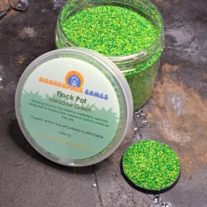 May include: A jar of Meadow Green flocking material for tabletop gaming, with a lid displaying the Moonmapper Games logo and product details. A small, round base covered in the green flocking is also present.