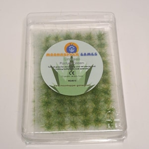 Può includere: Un contenitore di plastica con ciuffi di erba verde per il modellismo ferroviario o i giochi in miniatura. Il contenitore ha un'etichetta che recita "Grasses Pasture Green" e "Moonhopper Games".