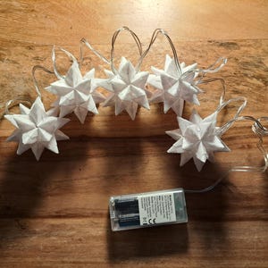 Op de afbeelding: Een reeks witte stervormige lichtslingers met een batterijpack. De lichten zijn gerangschikt op een houten oppervlak. De sterren hebben een textuur, origami-achtige uitstraling en de draad is transparant.