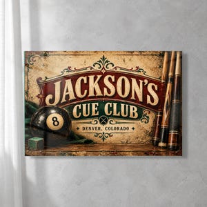 May include: A rectangular wall sign featuring the text Jackson's Cue Club Denver, Colorado. The design includes a black 8-ball, a small green cube, and three wooden pool cues against a weathered, vintage-style background with ornate borders.