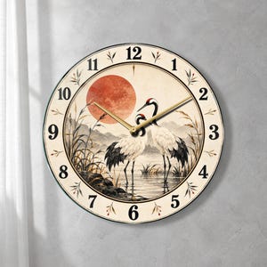 May include: A round wall clock featuring a cream-colored face with black numerals. The center displays an illustration of two white cranes with red crowns standing in water near reeds, set against a backdrop of mountains and a large red sun.
