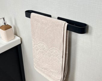 May include: A black towel bar holds a beige towel with a decorative, embroidered edge. The towel is hanging in a bathroom setting. A soap dispenser is visible in the background.