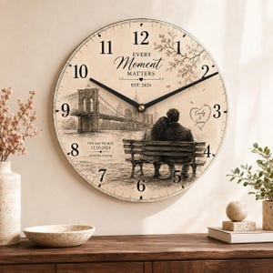 May include: A round wall clock with a beige face featuring a black sketch of a couple sitting on a bench facing the Brooklyn Bridge. The clock displays the text Every Moment Matters, Est. 2024, The Day We Met 12.10.2024, Austin, Texas, and Emily & James.