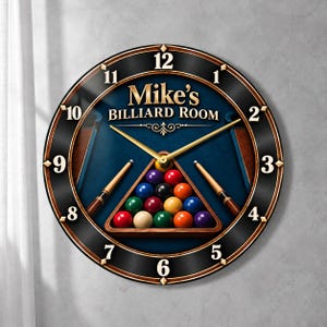May include: A round wall clock with a black and wood-toned frame, featuring the text Mike's BILLIARD ROOM. The clock face displays a blue pool table surface, a triangular rack of colorful billiard balls, and two crossed pool cues.