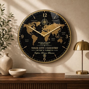 Op de afbeelding: Een ronde wandklok met een zwarte wijzerplaat, een gouden wereldkaart, gouden wijzers en gouden cijfers op 3, 6, 9 en 12 uur. De tekst bevat "YOUR CITY / COUNTRY", "40.7128 N, 74.0060 W", "SOUTIER ADVENTURE BEGINS HERE" en "Explore. Dream. Discover."