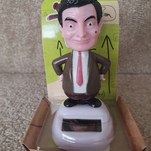 May include: A solar-powered Mr. Bean bobblehead figure. The figure is wearing a brown suit with a pink tie and black shoes. The base is white with a solar panel. The packaging is cardboard with green accents.