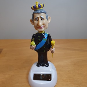 以下が含まれることがあります： 王室の制服を着て王冠をかぶった男性の首振り人形。 金色のトリムが付いた黒いジャケット、青いサッシュを着て、笏とオーブを持っています。 ベースはソーラーパネル付きの白です。