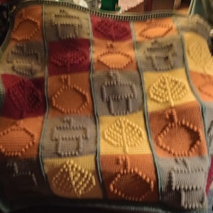 May include: A colorful, handmade knitted blanket with a repeating pattern of geometric shapes. The blanket features squares in shades of orange, yellow, red, and gray, with each square containing a raised design. The overall design is a patchwork style.