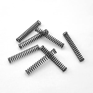 May include: A collection of small, black coil springs scattered on a white surface. The springs are tightly wound and have a shiny, metallic appearance. The image is a close-up, highlighting the details of the springs.