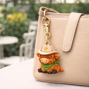 May include: A Highland Cow keychain with a 3D model design. The keychain features a brown cow wearing a tan cowboy hat and a green scarf. The keychain is attached to a gold-colored clasp and chain, hanging from a beige purse.