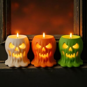 May include: Three Halloween skull candles in white, orange, and green. Each candle has a lit wick and a carved skull face with glowing eyes and mouth. The candles are on a dark surface with a blurred background.