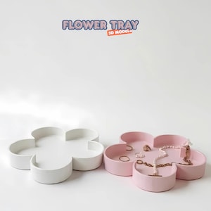 May include: Two flower-shaped trays. One is white, the other is pink and holds jewellery. The trays have four curved sections. The text "FLOWER TRAY 3D MODEL" is at the top. The trays are on a white surface.