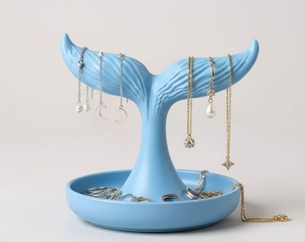 May include: A light blue whale tail jewelry holder. The holder has a round base and a tail-shaped top for hanging necklaces. Several necklaces and rings are displayed on the holder. The jewelry holder is made of a smooth, solid material.