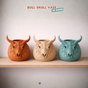 Puede incluir: Tres jarrones con forma de calavera de toro en naranja, beige y turquesa, cada uno con intrincados dise&ntilde;os florales y cuernos curvos. Los jarrones se exhiben en una estanter&iacute;a de madera sobre un fondo blanco. El texto "BULL SKULL VASE 3D MODEL" est&aacute; en la parte superior.