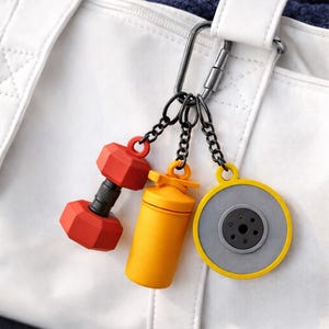 Puede incluir: Una bolsa de gimnasio blanca con un mosquetón plateado que sostiene tres llaveros con temática de fitness. Los llaveros incluyen una mancuerna roja, una botella mezcladora amarilla y una placa de pesas gris y amarilla.