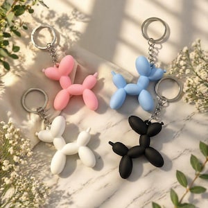 Puede incluir: Cuatro llaveros de perro globo en rosa, azul, blanco y negro. Cada llavero tiene una cadena y un anillo plateados. Las figuras de perro est&aacute;n hechas de un material liso y est&aacute;n dispuestas sobre una superficie clara con vegetaci&oacute;n.