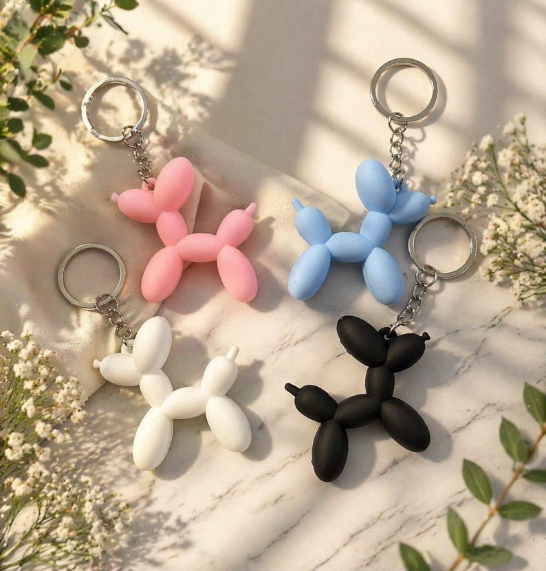 Puede incluir: Cuatro llaveros de perro globo en rosa, azul, blanco y negro. Cada llavero tiene una cadena y un anillo plateados. Las figuras de perro est&aacute;n hechas de un material liso y est&aacute;n dispuestas sobre una superficie clara con vegetaci&oacute;n.