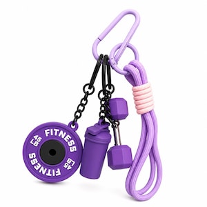 Puede incluir: Un llavero morado con temática de fitness que presenta un peso de barra, una botella mezcladora y una mancuerna. El peso de la barra está etiquetado como "45 LB" y "FITNESS". El llavero incluye un mosquetón morado y una cuerda morada y rosa.