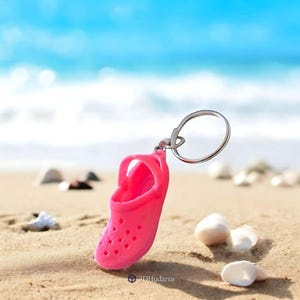 May include: A pink rubber shoe keychain with a silver key ring. The keychain is on a sandy beach with seashells.