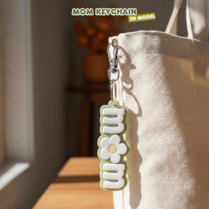 May include: A green and white keychain with the word "mom" in a flower design. The keychain is attached to a silver clasp and ring. The text "MOM KEYCHAIN" and "3D MODEL" are in the background.