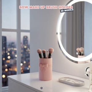 May include: A pink cylindrical makeup brush holder with a bow detail, holding several makeup brushes. The text "BOW MAKE UP BRUSH HOLDER" is at the top of the image. The holder is on a white surface near a lighted mirror.