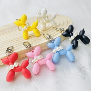 Puede incluir: Una colecci&oacute;n de llaveros de perro globo de colores: rojo, rosa, amarillo, azul, blanco y negro. Cada perro est&aacute; adornado con un collar de perlas y un cierre de llavero plateado. Los llaveros se exhiben en una bandeja de madera.