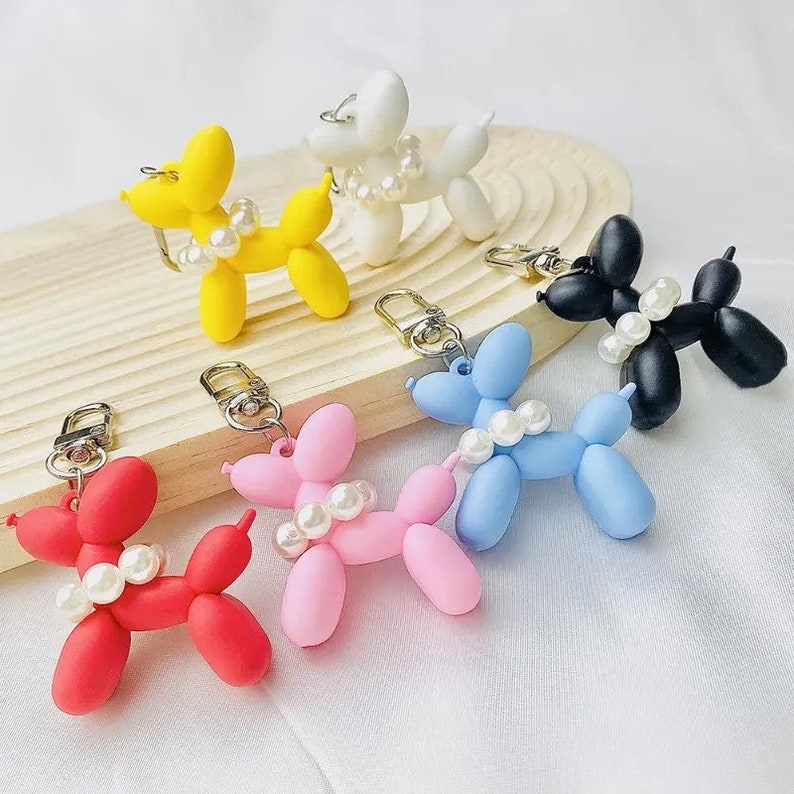 Puede incluir: Una colecci&oacute;n de llaveros de perro globo de colores: rojo, rosa, amarillo, azul, blanco y negro. Cada perro est&aacute; adornado con un collar de perlas y un cierre de llavero plateado. Los llaveros se exhiben en una bandeja de madera.