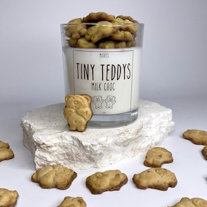 May include: A glass jar filled with milk chocolate teddy bear biscuits. The jar has a label that reads "Milky's Tiny Teddys Milk Choc". There are several biscuits scattered around the jar on a white surface.