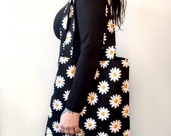 May include: Black tote bag with a daisy pattern. The bag features white daisies with yellow centers on a black background. The tote bag is being carried over the shoulder.